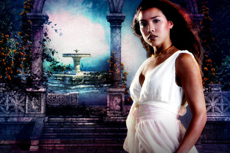 A picture of Maricela (tall, brunette, latina, dressed in a white dress) in front of a fountain behind two stone arches in the center of a garden maze.
